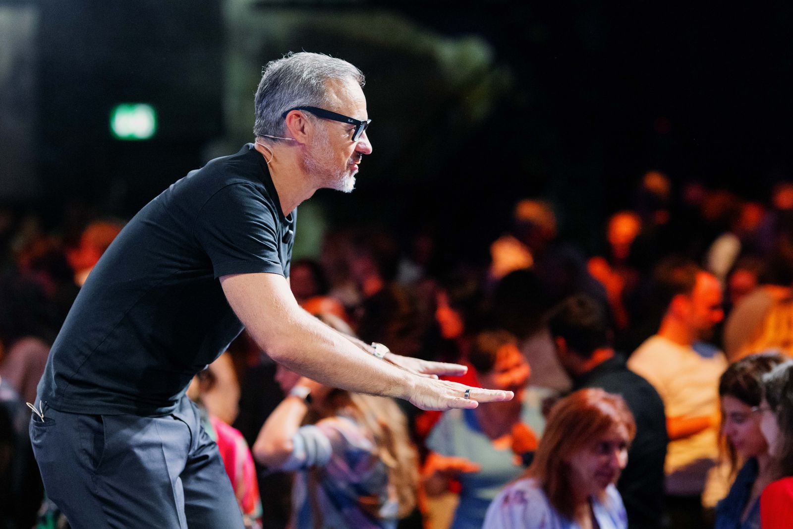 Eric Edmeades in black-framed glasses and a black t-shirt, leaning forward with both arms out toward a crowd.