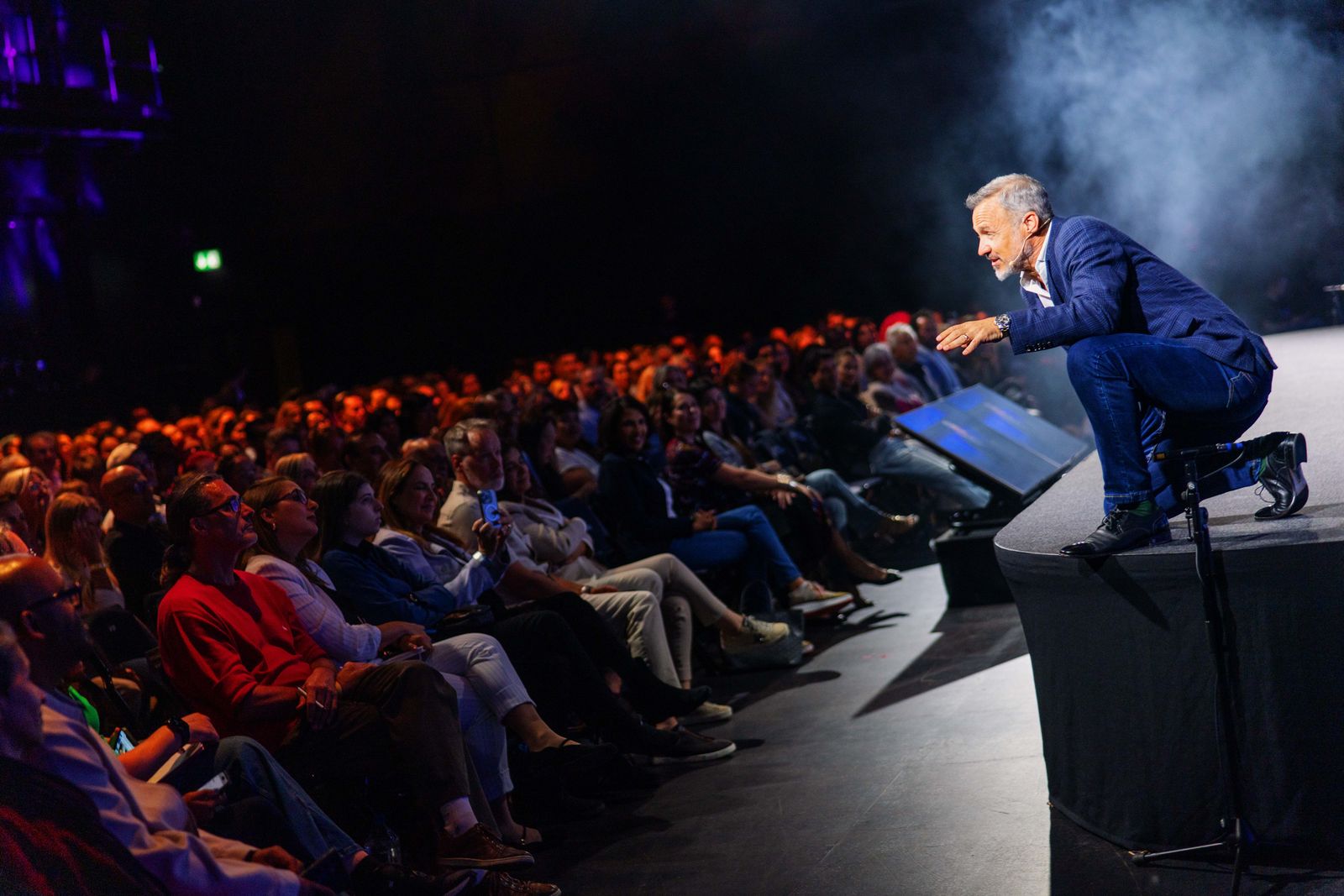 Eric Edmeades in a navy suit crouching at the edge of a raised stage, arms extended toward a seated audience in a dim auditorium.
