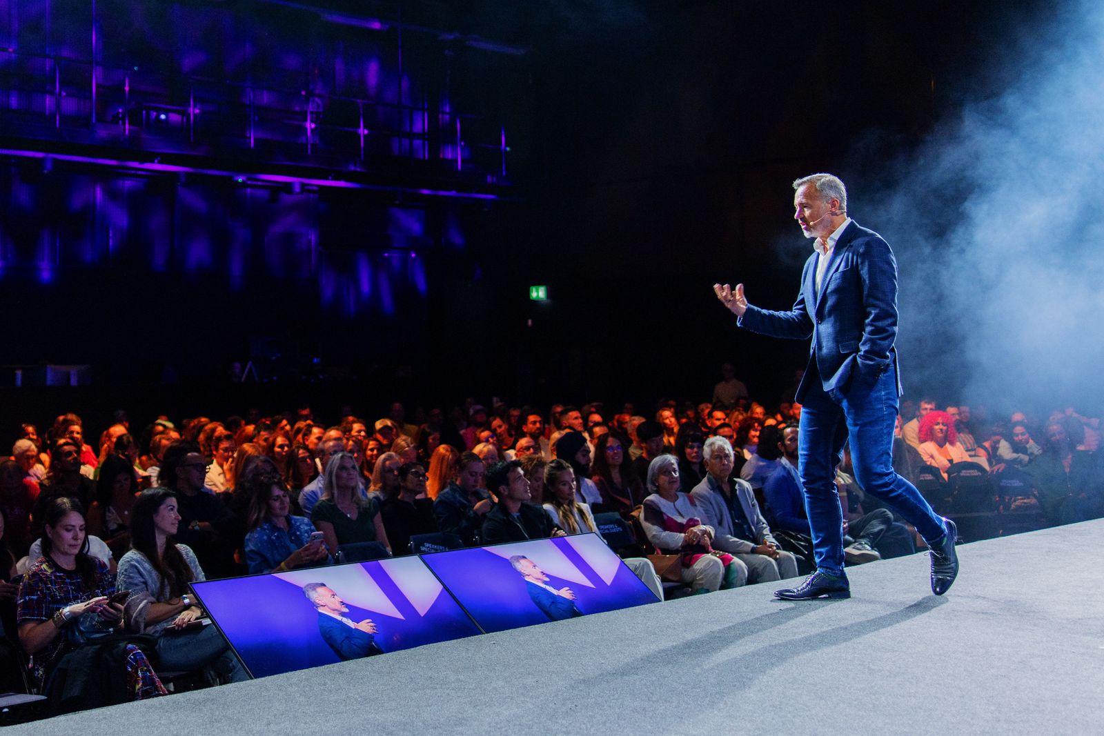 Eric Edmeades in a navy suit walking across a lit stage and gesturing, with a seated audience and three screens showing a close-up of him.