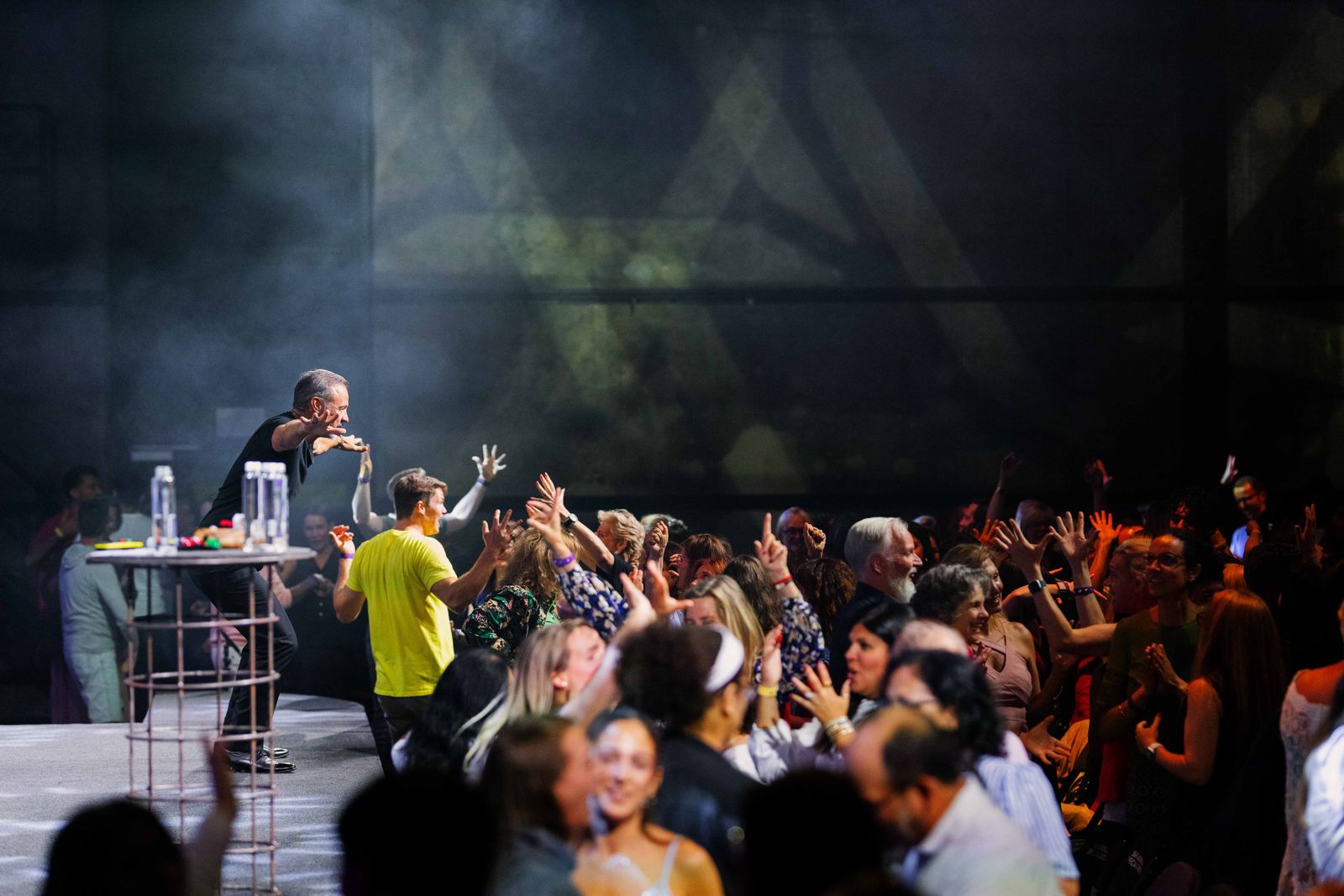 Eric Edmeades on a raised stage at the left, reaching toward a dense crowd with many raised hands, in stage haze.